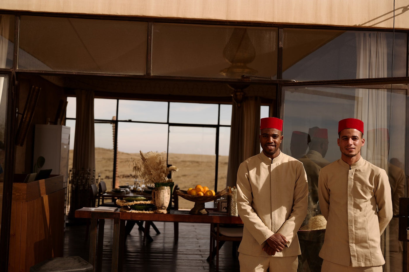 Two waiters in beige uniforms and red fez standing at the door of a tented lounge