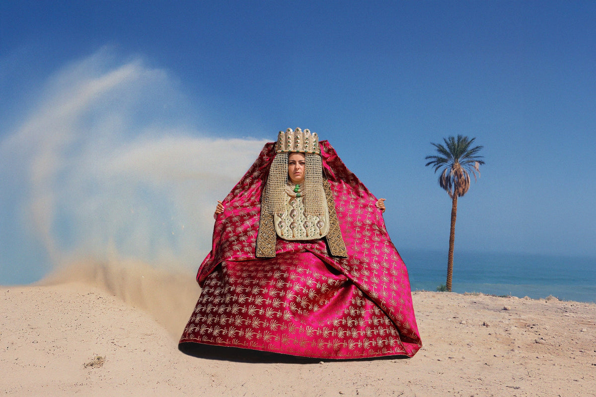 Woman in an embroidered fuchsia caftan and headdress on a beach, sand blowing behind her