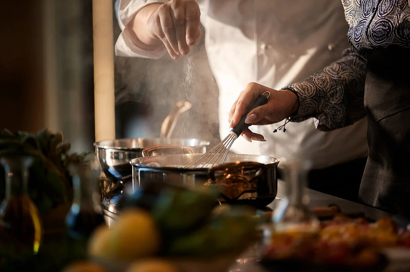 Chef in a dark jacket whisking a sauce in a steaming pan, ingredients in the foreground