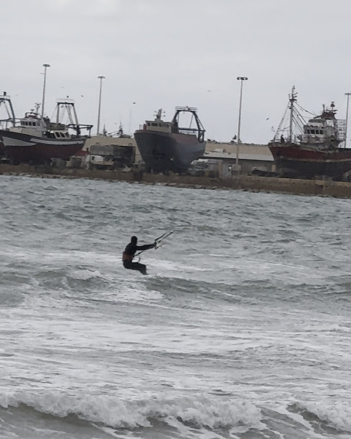 Un kitesurfeur dans les vagues de la baie d'Essaouira, des chalutiers amarrés au port derrière