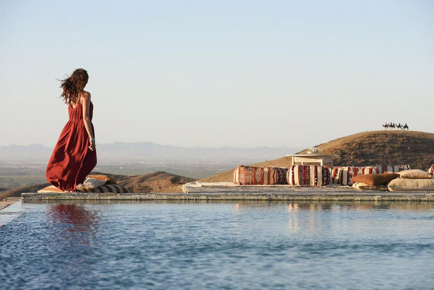Woman in a red dress walking along an infinity pool above a stone desert at golden hour, camels on a ridge