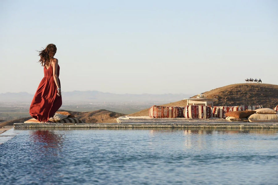 Woman in a red dress walking along an infinity pool above a stone desert at golden hour, camels on a ridge