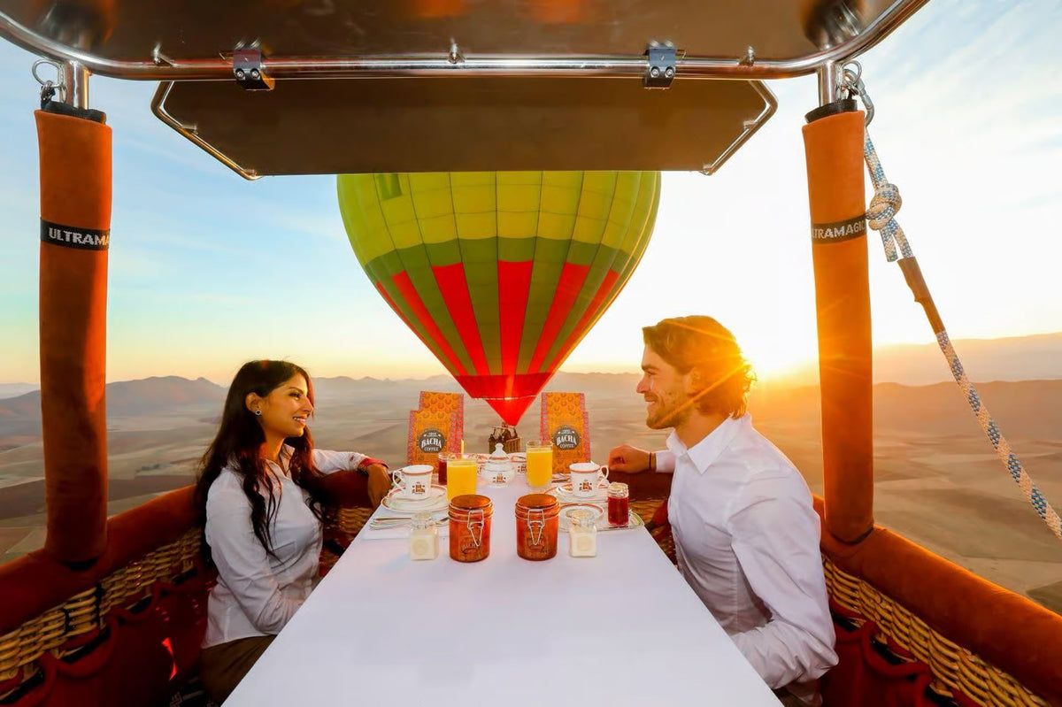 Couple seated at a breakfast table inside a hot-air balloon basket at sunrise, second balloon behind