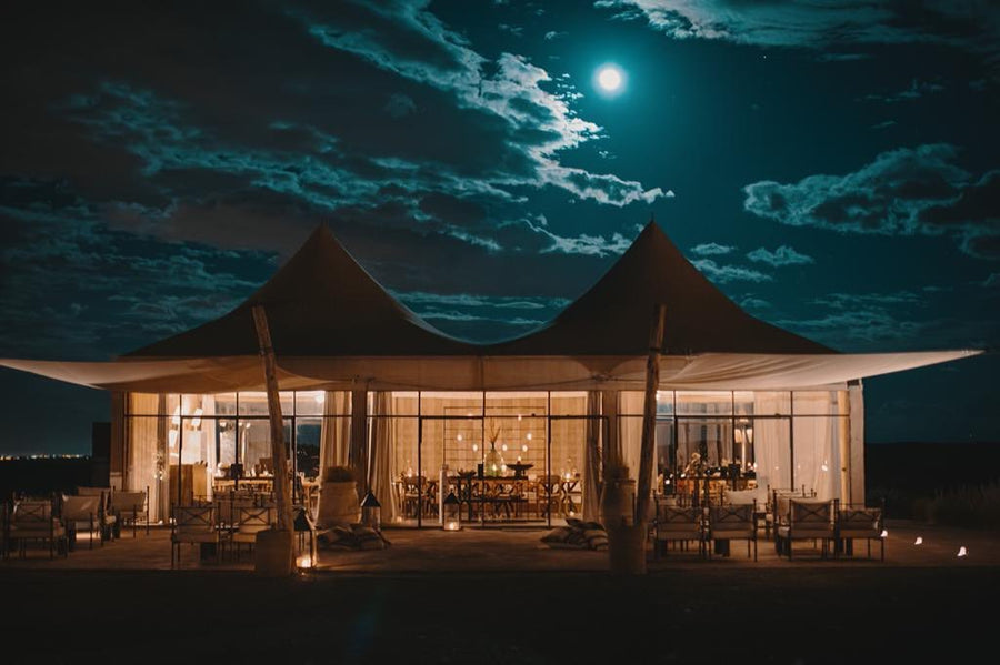 Glass-walled tented lodge lit from within under a full moon and clouds