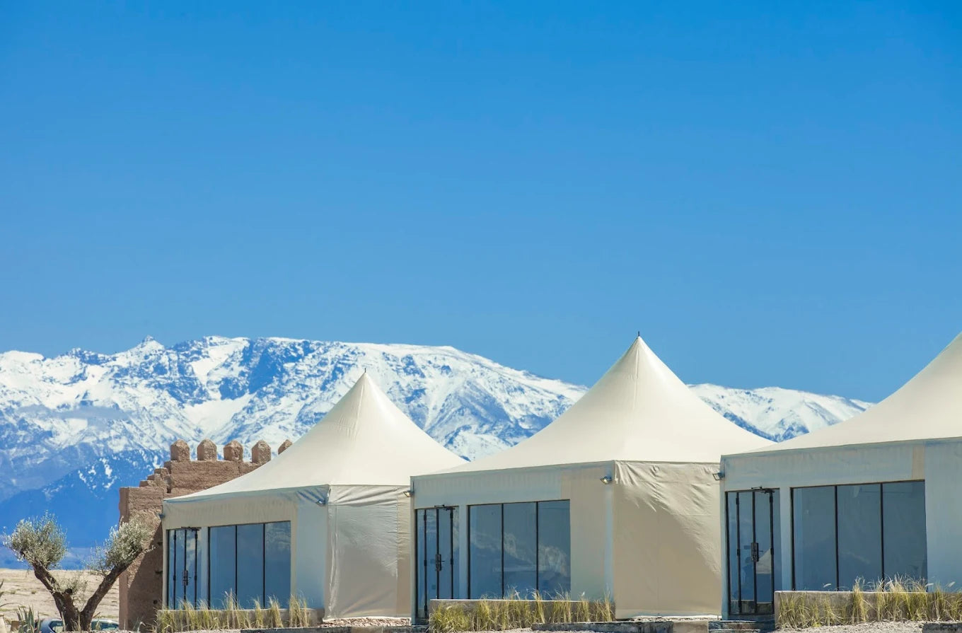 White canvas lodges with glass fronts under a clear sky, snow-capped Atlas mountains behind