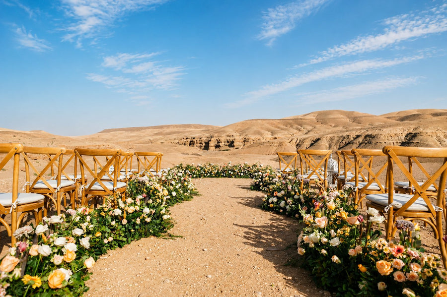 Wooden chairs and rows of pale roses lining an aisle in a stone desert under a blue sky