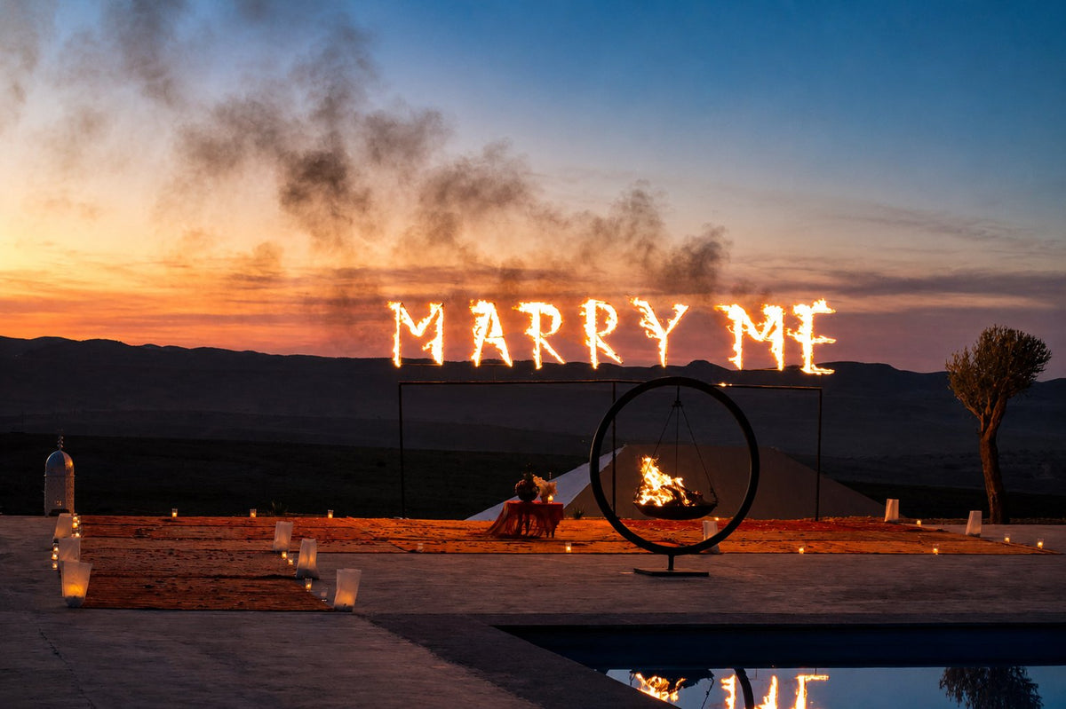 Burning MARRY ME letters and a ring of fire at sunset above a pool, candles on the ground