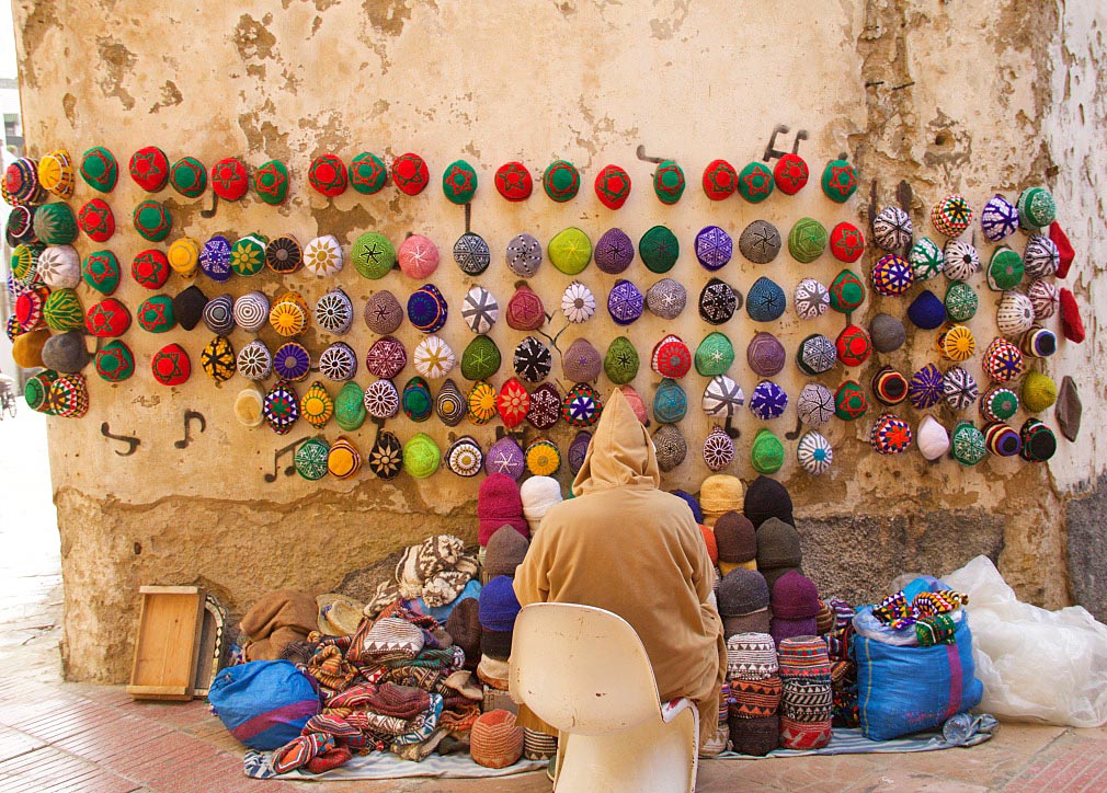 Colourful knitted hats hung on a weathered wall above a seated vendor