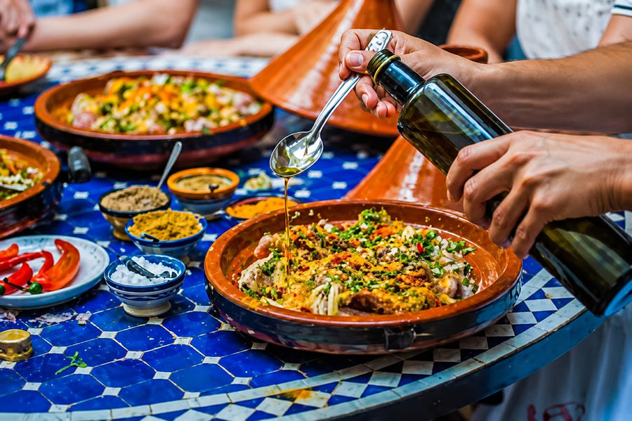 Olive oil poured over a tagine on a blue zellige table, side dishes around