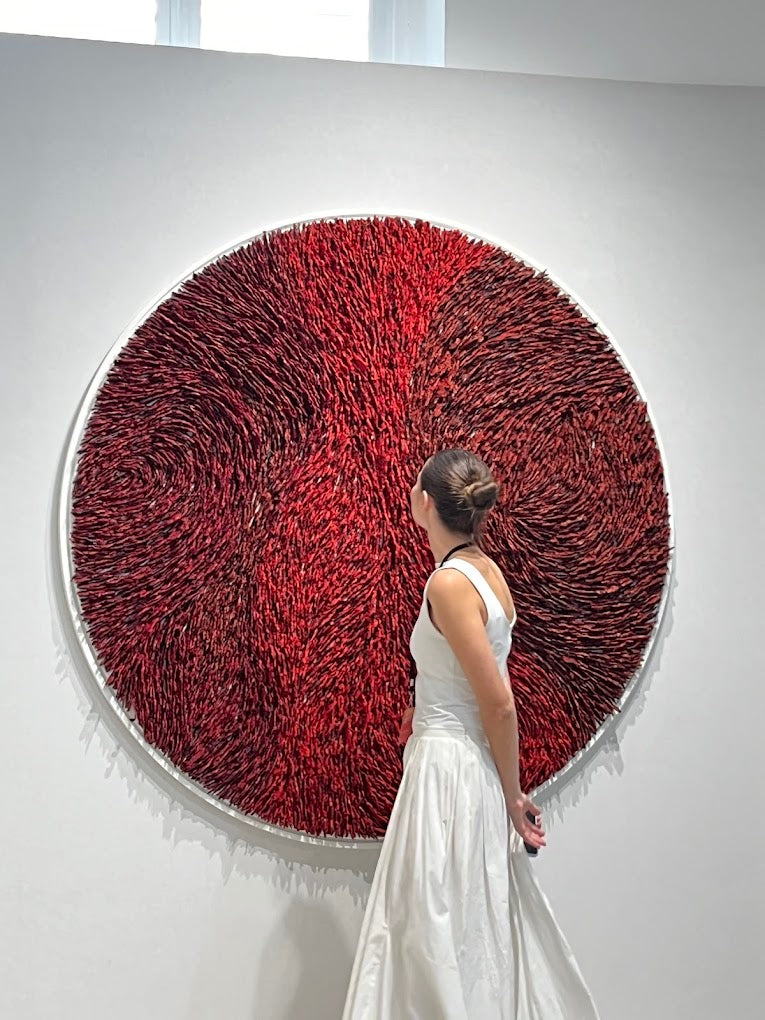 Woman in a white dress viewing a large circular red textile artwork in a white gallery