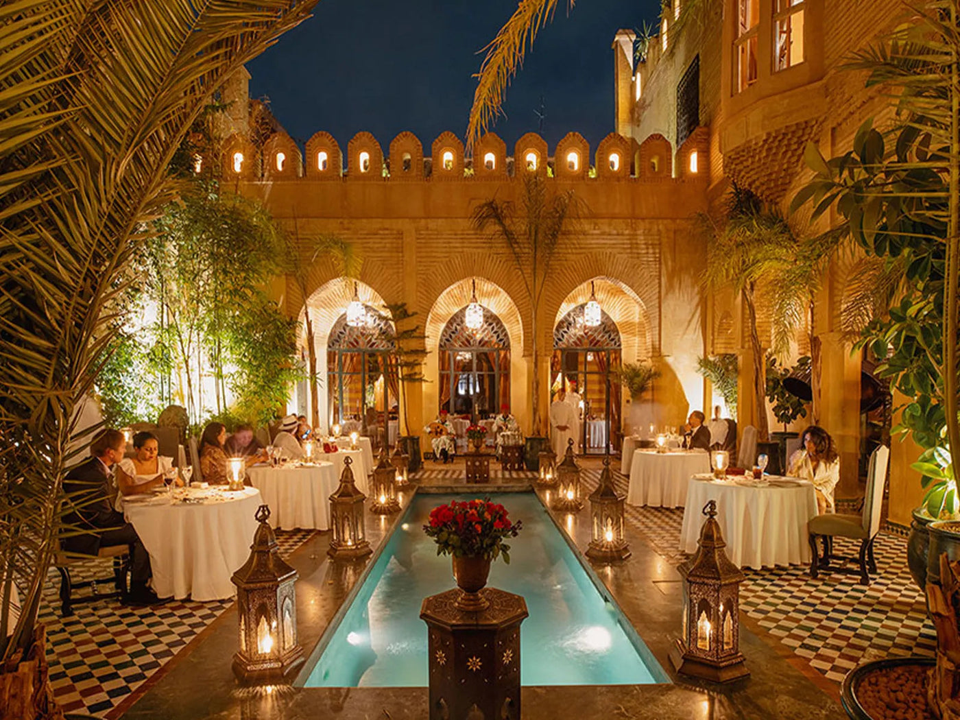 Riad courtyard set for dinner at night, lanterns around a long pool between arcades and palms