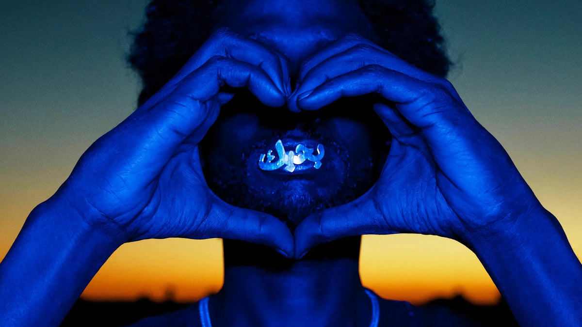 Hands painted blue forming a heart around Arabic calligraphy against a sunset sky