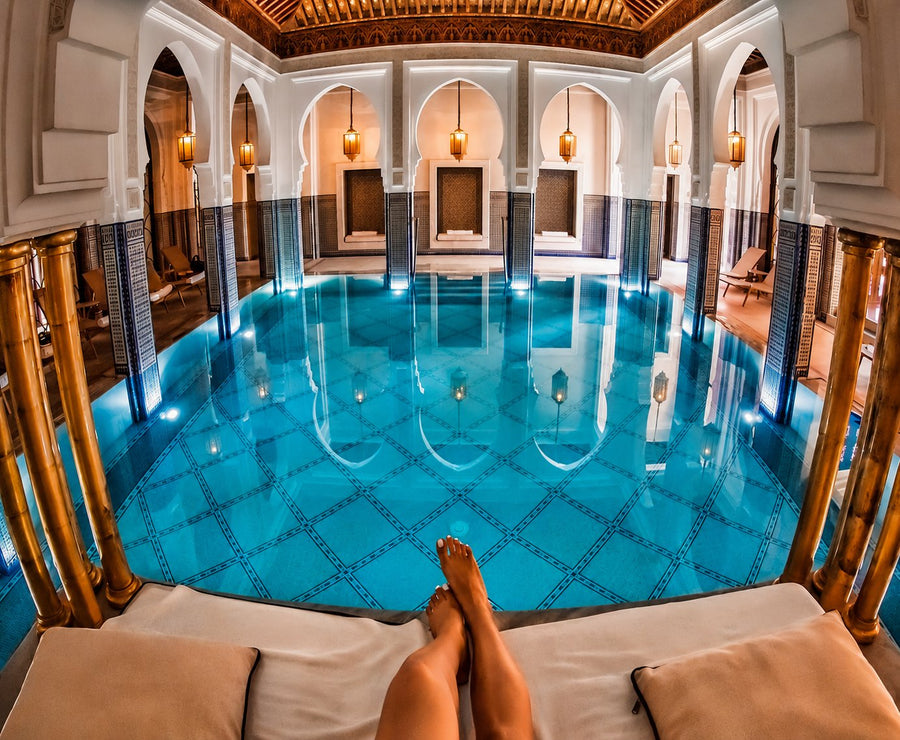 Indoor pool under carved white arches and hanging lanterns, bare feet resting on a lounger in the foreground