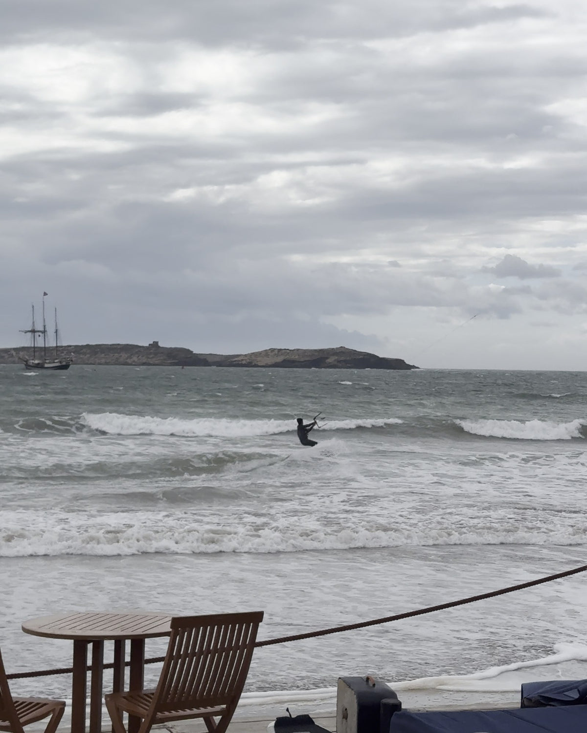 Une table et des chaises face à la baie d'Essaouira, un kitesurfeur et l'île au large sous un ciel gris