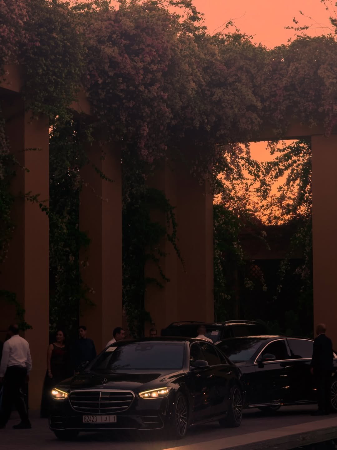 Black Mercedes sedan with headlights on at a hotel entrance at dusk, staff waiting